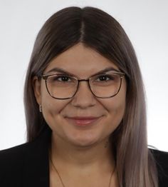 Eine lächelnde Person mit Brille und langen Haaren vor einem neutralen Hintergrund.