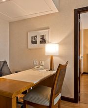 Escritório em hotel com mesa, cadeira e lâmpada; quarto visível ao fundo.