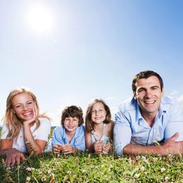 Family lying on grass under a bright sunny sky, all smiling at the camera.