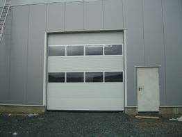 Graues Industrietor mit Fenstern neben einer kleinen weißen Tür, beide vor einer grauen Wand.