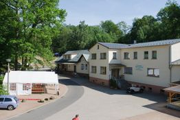 Gebäude in einer ländlichen Umgebung mit Wald im Hintergrund und einem Zelt im Vordergrund.