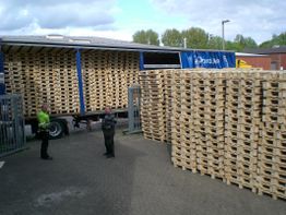 LKW liefert Paletten, während zwei Personen daneben stehen, großer Stapel Paletten im Hintergrund.