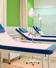 Camillas en una sala de fisioterapia con luces y cortinas, paredes verdes y blancas.