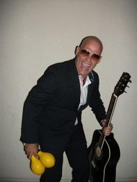 Hombre con gafas de sol, traje oscuro, guitarra y maracas amarillas, sonriendo ampliamente.