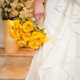 Bride holding a bouquet of yellow roses, standing on stairs in a flowing white wedding dress.