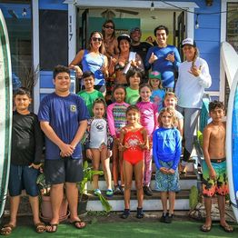 Group of children and adults posing on steps of a blue building, wearing casual and swimwear.