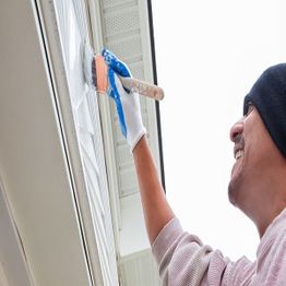 Person smiling while painting a house exterior with a brush and wearing a beanie.