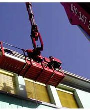 Roter Kran hebt Arbeitsplattform vor einem Gebäude hoch, Person hinter gelbem Fensterrollo sichtbar.