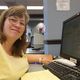 Woman in a yellow shirt smiling at a desk with a computer and notepad.