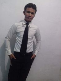 Homem em pé contra parede branca, vestindo camisa branca e gravata preta, com mãos nos bolsos.