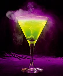 Leuchtend grüner Cocktail in Martini-Glas mit dampfendem Rauch vor lila Hintergrund.