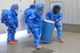 People in blue hazmat suits carry a blue barrel inside a building.