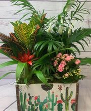 Caja decorativa con plantas verdes y flores rosadas sobre fondo de madera blanca.