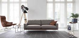 Modernes Wohnzimmer mit grauem Sofa, Lederstuhl, Stehlampe und großem Fenster im Hintergrund.