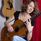 Eine Frau spielt Gitarre vor einem roten Vorhang, im Hintergrund hängen Gitarren an der Wand.