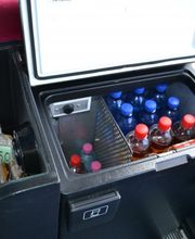 Kleiner Kühlschrank mit Wasser- und Saftflaschen in einem Fahrzeug, daneben ein Korb mit Snacks.