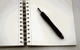 Offenes Notizbuch mit Spiralbindung und schwarzem Stift auf einer weißen Oberfläche.