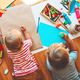 Children drawing on paper with crayons and craft supplies scattered on the floor.