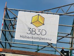 Gerüst mit Banner "3B3D Höhenkompetenz" und einem gelben Würfellogo, blauer Himmel im Hintergrund.