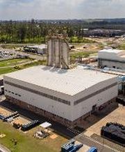 Fábrica em área industrial com vegetação ao fundo e céu parcialmente nublado.