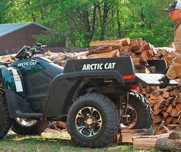 Hombre cargando leña en un vehículo todo terreno Arctic Cat en un campo con árboles.
