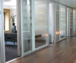 Büroflur mit Glaswänden, Holzboden und Blick in ein Zimmer mit Sesseln und Pflanze.
