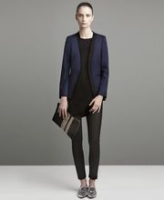 Modella in piedi con giacca blu, pantaloni neri e borsa, su sfondo grigio neutro.