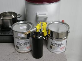 Dosen mit Korrosionsschutzfett, Heizplatte, Topf und Sprühgerät auf Tisch.