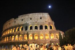 Colosseo illuminato di notte con la luna e folla di persone in movimento.