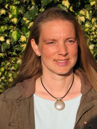 Eine Frau mit langen Haaren lächelt im Sonnenlicht, im Hintergrund grüne Blätter.