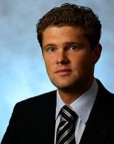 Ein Mann in Anzug mit Krawatte, vor blauem Hintergrund, schaut in die Kamera.