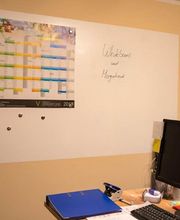 Whiteboard mit Magneten, links Wandkalender. Vorne Schreibtisch mit Monitor und Ordner.