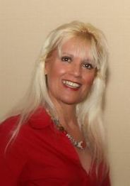 Eine lächelnde Frau mit blonden Haaren trägt ein rotes Hemd vor einem beigen Hintergrund.