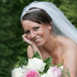 Braut mit Schleier und Diadem lächelt, stützt Kopf auf Hand, Blumenstrauß im Vordergrund.