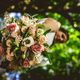 Bruid met boeket van roze en witte bloemen, kijkend door groene bladeren in buitenomgeving.