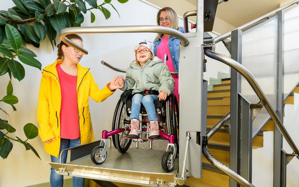 Ein Mädchen im Rollstuhl wird von ihrer Mutter auf den Homelifte, die Treppen hoch transportiert.