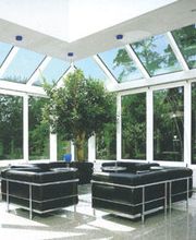Moderner Wintergarten mit Glasdach, schwarzen Sofas und grünem Baum, umgeben von viel Tageslicht.