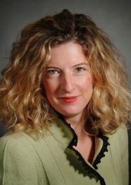 Eine Frau mit lockigem, blondem Haar und grüner Jacke lächelt vor neutralem Hintergrund.