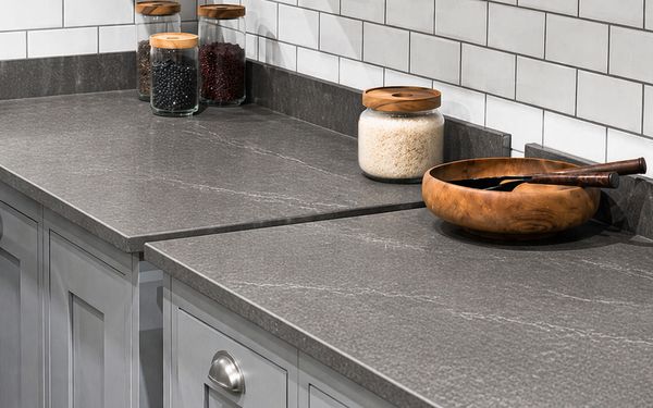Encimera de cocina de cerámica en tono gris con vetas sutiles, superficie mate y uniforme, acompañada de tarros de vidrio con alimentos y un bol de madera sobre muebles blancos y salpicadero de azulejos.