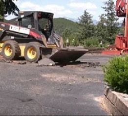 Skid steer loader breaking asphalt on a road with trees and hills in the background.