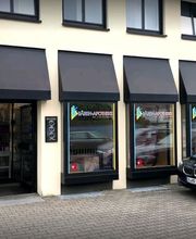 Gebäude mit Apothekenzeichen, Schaufenstern und einem geparkten schwarzen Auto davor.