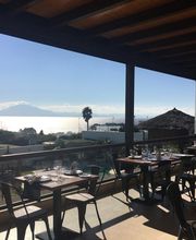 Restaurante con vista al mar y montaña bajo un cielo despejado y soleado.