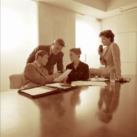 Vier Personen in einem Büro sprechen und betrachten Dokumente an einem Tisch.