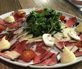 Carpaccio mit Rucola, Kirschtomaten, Parmesan, Pilzen und Cremetupfen auf einem Teller.