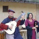 Zwei Musiker in mittelalterlichen Kostümen spielen Gitarre und Flöte vor einem Strohdachhaus.