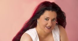 Eine Frau mit dunklem Haar und weißem Top vor rosa Hintergrund lächelt in die Kamera.