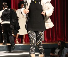 Person auf Laufsteg mit Sonnenbrille, Felljacke, gemusterten Leggings vor rotem Vorhang.