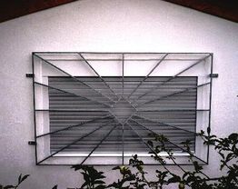 Fenstergitter mit geometrischem Design an weißer Wand, verdeckt von Pflanzen im Vordergrund.