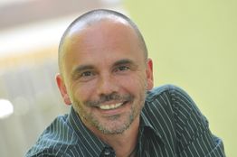 Ein lächelnder Mann mit Glatze und Bart trägt ein gestreiftes Hemd vor unscharfem Hintergrund.