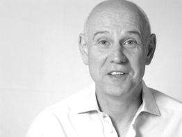 Schwarzweißfoto eines lächelnden Mannes mit Glatze im Hemd vor weißem Hintergrund.